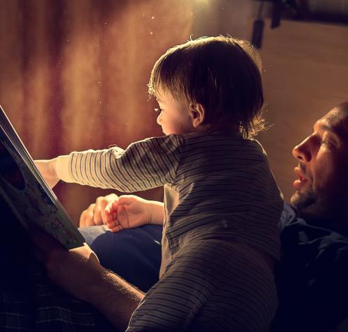 Fotografía de un hombre leyendo a un niño pequeño en su regazo. El adulto sostiene el libro abierto mientras el bebé estira su brazo hacia las páginas. Una luz cálida ilumina sus rostros contra un fondo oscuro, creando un ambiente íntimo de aprendizaje y vínculo.