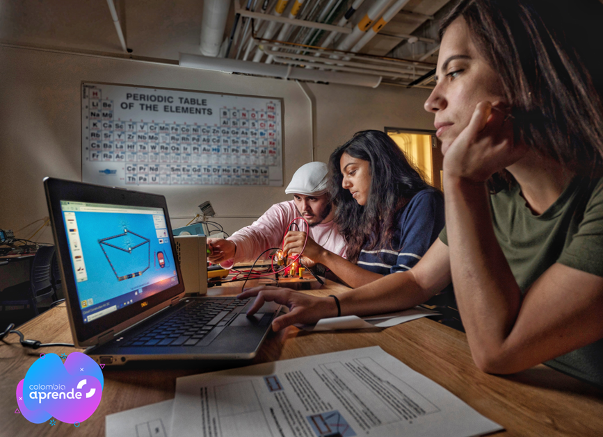 Dos estudiantes mujeres y un estudiante hombre trabajando en mesa frente a computador con simulaciones
