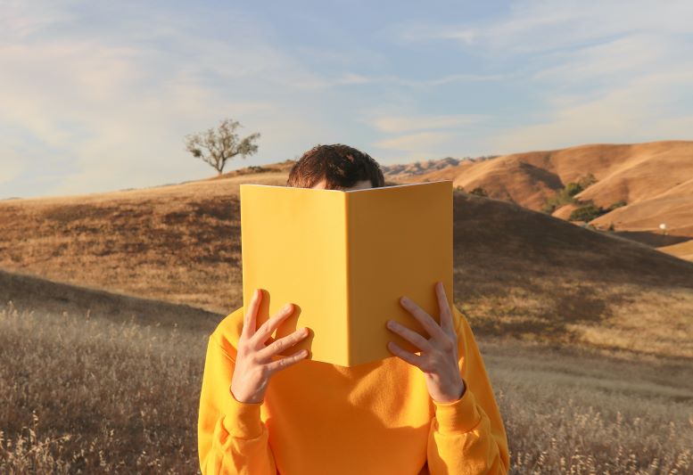 Joven vestido de amarillo, que tapa su cara con un libro amarillo que lee, con fondo de montañas