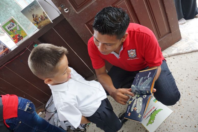 Profesor arrodillado junto a un niño pequeño le muestra imagenes de un libro ilustrado