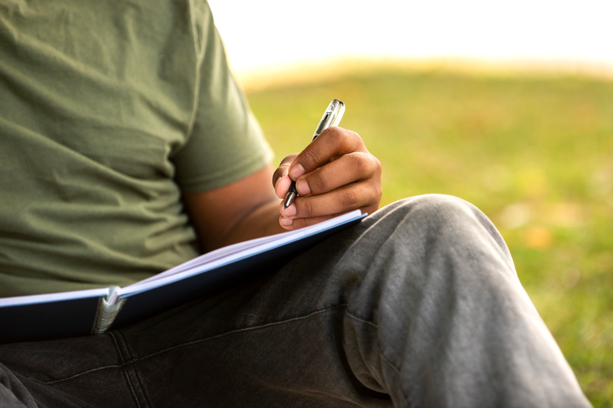 Foto de un estudiante de camiseta y blujean escribiendo en un cuaderno. 