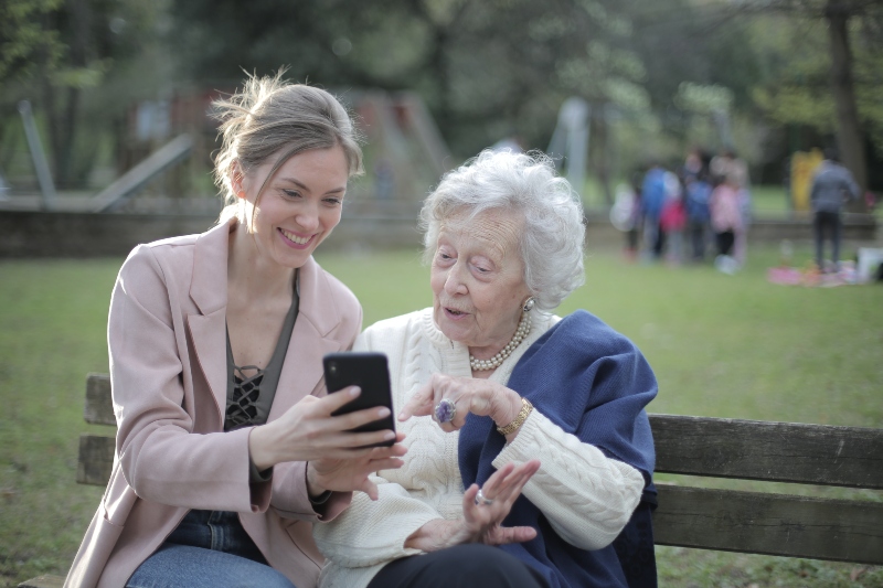 Foto de una mujer joven mostrándole la pantalla del celular a una mujer de pelo blanco mayor, mientras sonrien en un parque
