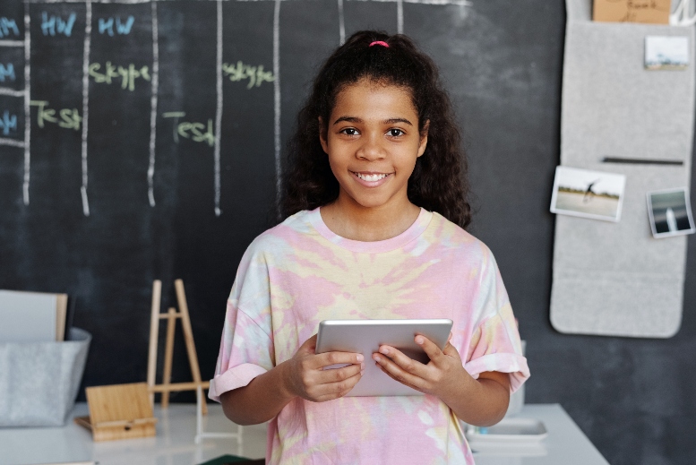 Adolescente de pelo negro largo, sonriente sostiene una tablet en sus manos y al fondo se ve un tablero verde y otros elementos escolares