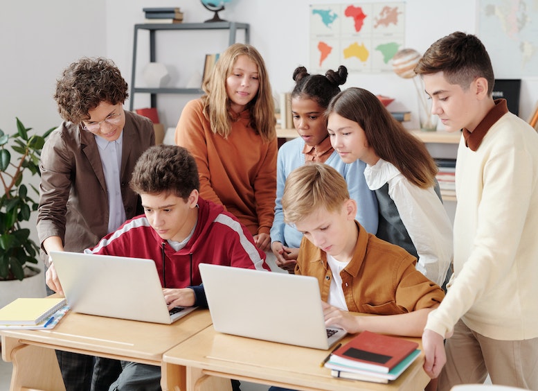 Cuatro adolescentes hombres y tres mujeres jóvenes, miran a dos de ellos que trabajan en un portátil cada uno, en un aula de clase