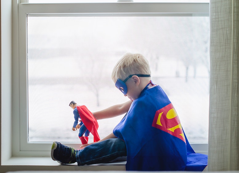 Foto de un niño disfrazado de Supermán y máscara, jugando con un muñeco de superman, contra una ventana