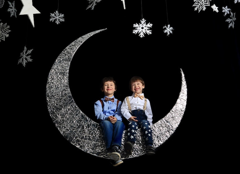 Dos niños sentados en una media luna plateada con fondo negro y estrellas plateadas