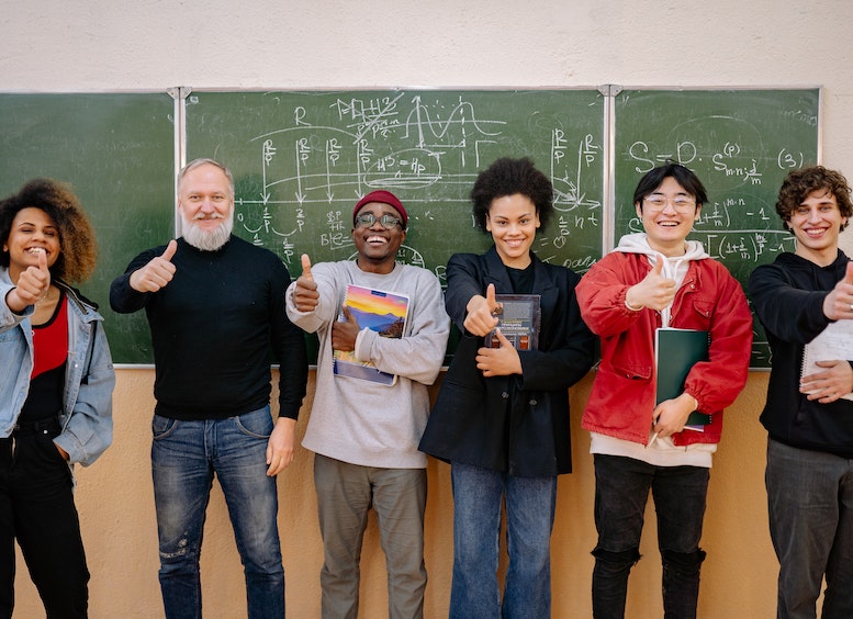 Dos mujeres y cuatro hombres adultos, parados adelante de un tablero de clase, sosteniendo libros y levantando el dedo pulgar en señal de aprobación