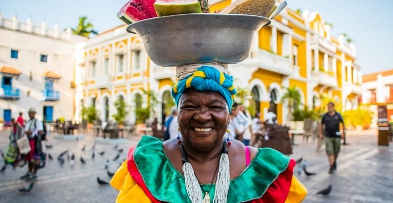 foto de una mujer palenquera con un platón con frutas sobre la cabeza y de fondo está el carnaval de Barranquilla