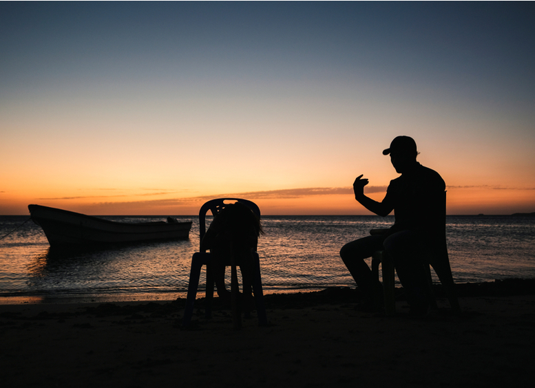 Hombre habla con su hijo sentado en la playa, con fondo de barco al atardecer en el  cabo de la Vela, Guajira, Colombia
