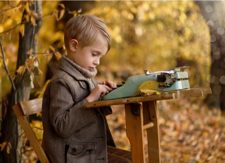 Niño rubio sentado en una silla con una máquina de escribir, en un bosque