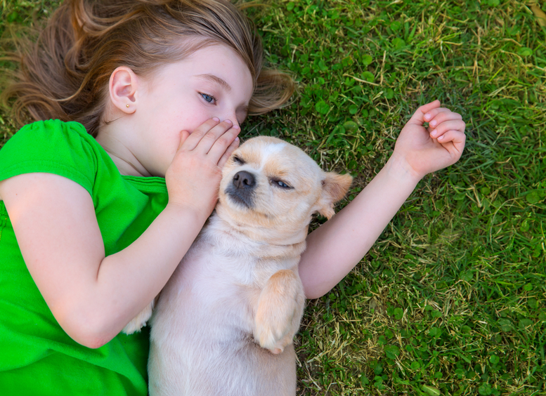 niña pelirroja, vestida de verde, tirada en el pasto junto a su perro chihuahua, a quien le dice un secreto