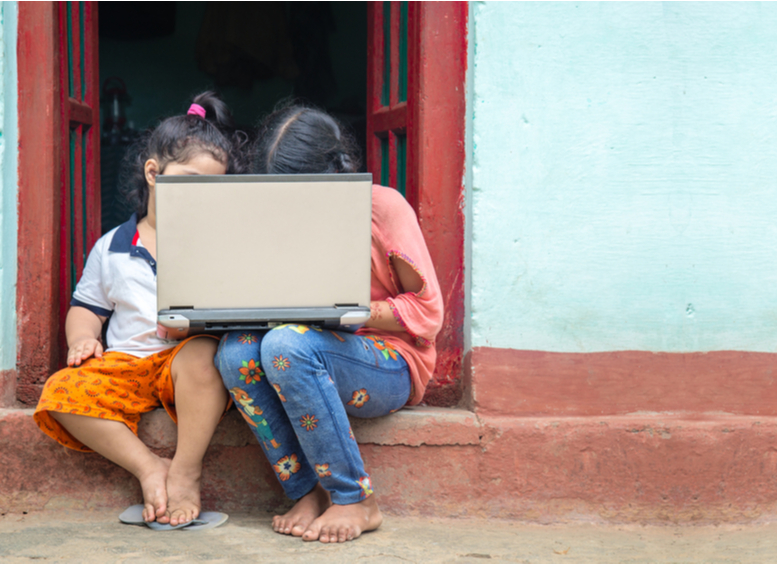 Dos niñas sentadas en la puerta de una casa, sostienen un computador en el regazo, y estudian