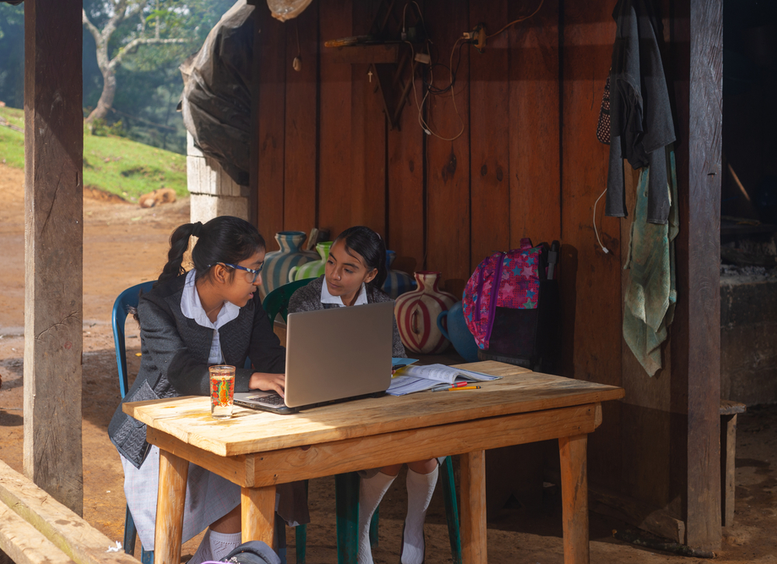 Dos niñas de zona rural, con uniforme, sentadas frente a una mesa con un computador, en las afueras de la casa con naturaleza al fondo