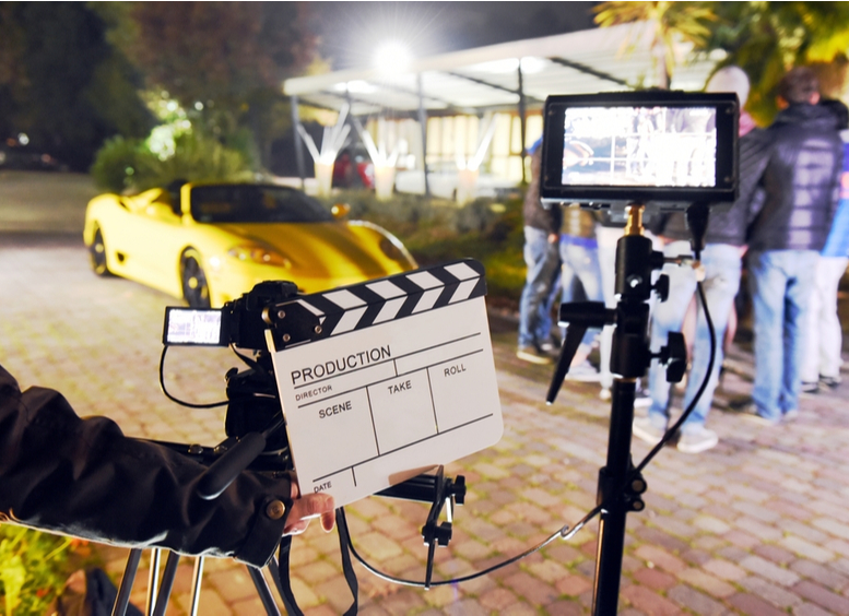 Operador sosteniendo tablero durante la producción de cortometraje al aire libre por la noche con automóvil deportivo amarillo y actor en el escenario. Enfoque en el tablero y monitores  