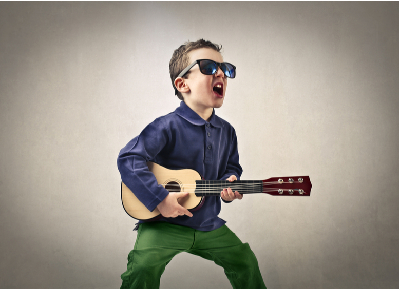 Niño con gafas oscuras, camisa azul y pantalón verde, con una guitarra tocando y cantando como estrella de rock