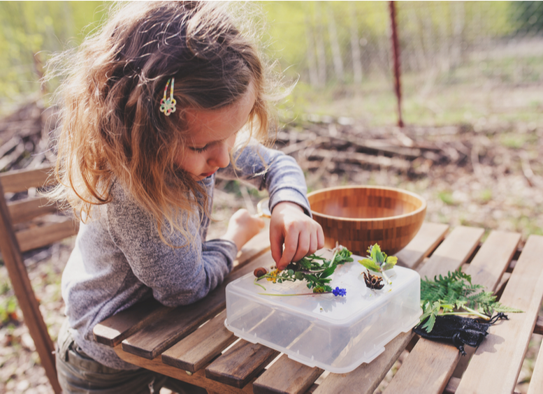 Foto de niña sentada en una mesa al aire libre, mirando las plantas que tiene encima 
