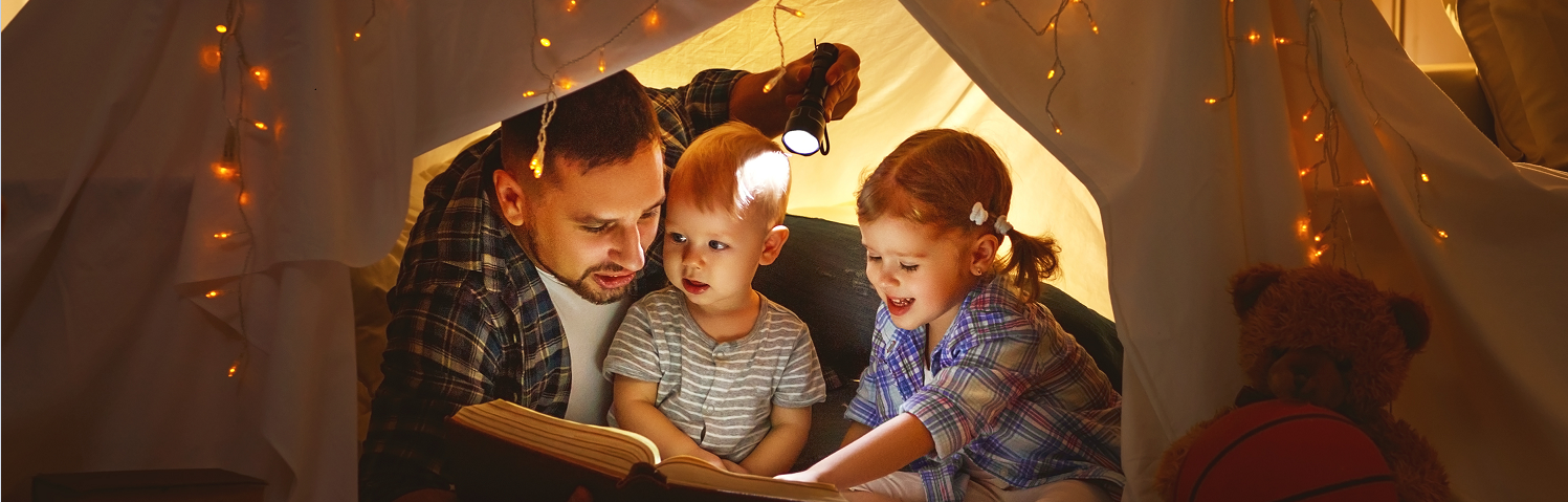 Familia de papa y dos hijos leyendo en una carpa con luces de navidad 
