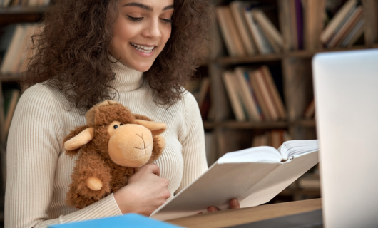 Una profesora joven, trigueña de pelo negro, sentada con una biblioteca de fondo, sostiene a una oveja cafe de juguete, y lee un libro de cuentos frente a una computadora portatil