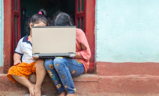 Dos niñas sentadas en la puerta de una casa, sostienen un computador en el regazo, y estudian