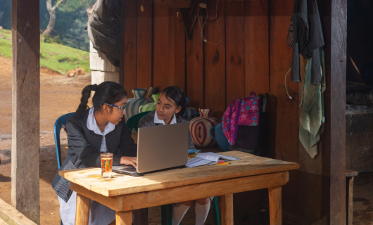 Dos niñas de zona rural, con uniforme, sentadas frente a una mesa con un computador, en las afueras de la casa con naturaleza al fondo