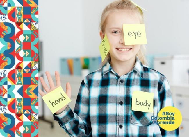 Foto de una niña con pegatinas de las partes del cuerpo en ingles que dice eye, body, hand