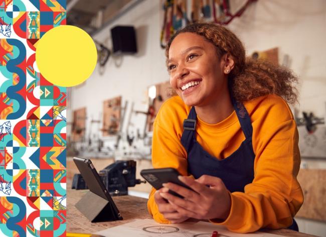 Foto de una mujer sonriendo, con celular en la mano y tablet sobre la mesa