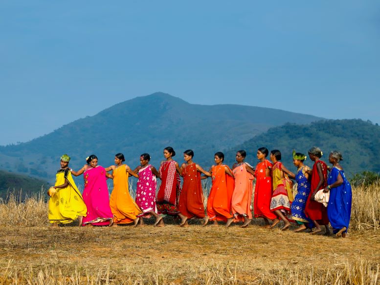 13 mujeres jovenes nativas con batas largas de colores caminan abrazadas, en el campo de día