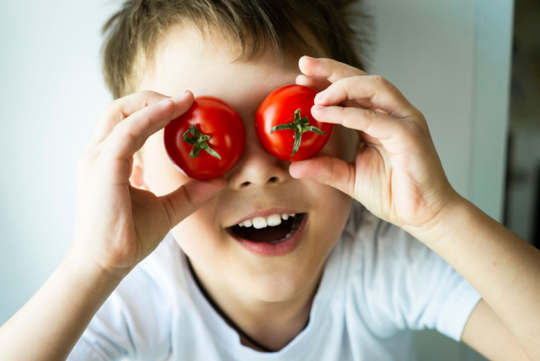 un niño alegre de entre 8 y 10 años de edad con un fondo claro sostiene tomates rojos en las manos y los pone en sus ojos