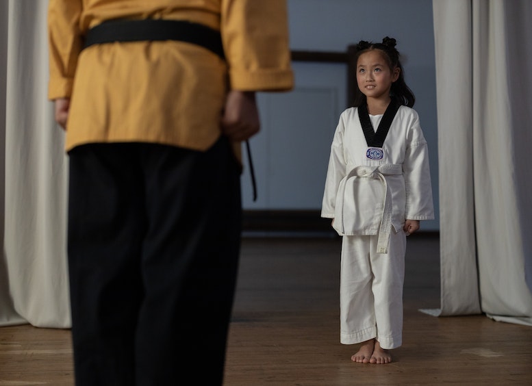 Niña con vestido blanco de artes marciales mira a su profesor que está de espalda a la cámara, con blusa amarilla y pantalón negro