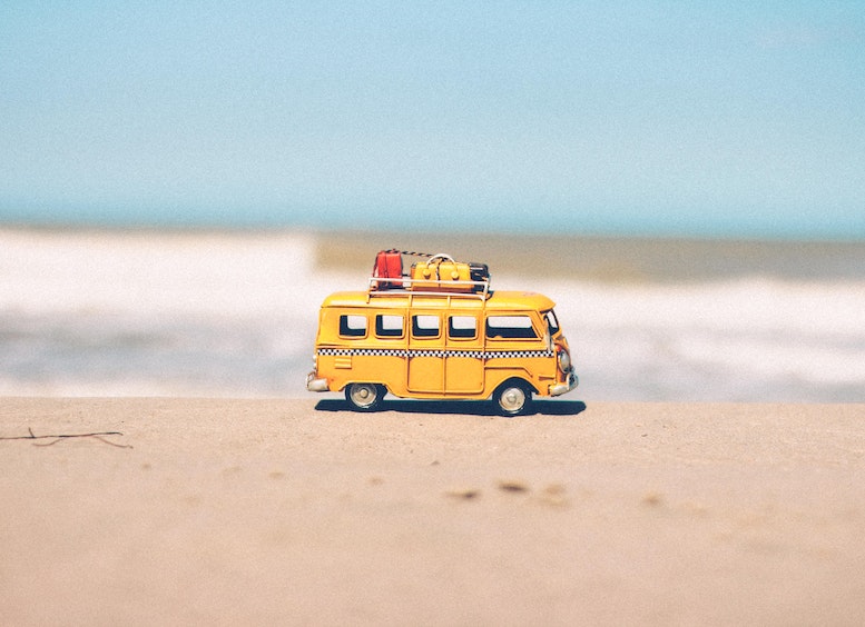 Foto de una furgoneta amarilla miniatura con maletas en su techo, sobre la arena y con el fondo del mar