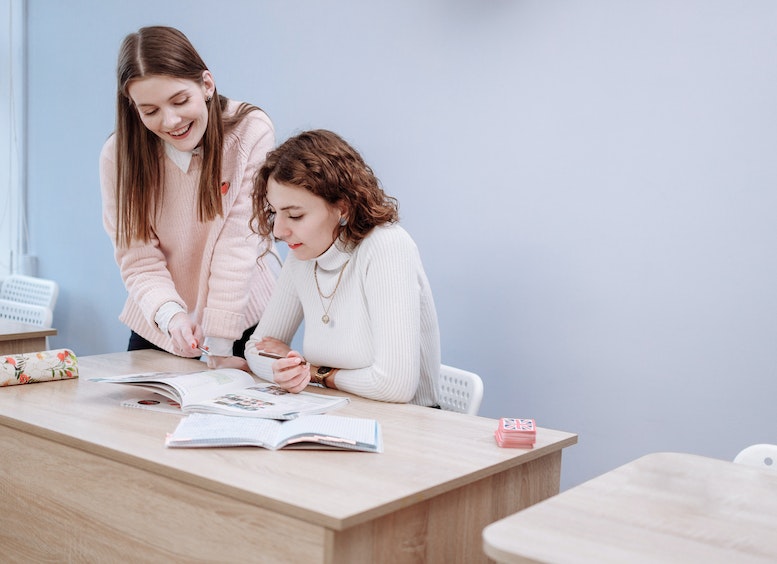 Alumna de pie le muestra cuaderno a maestra que está sentada en una mesa