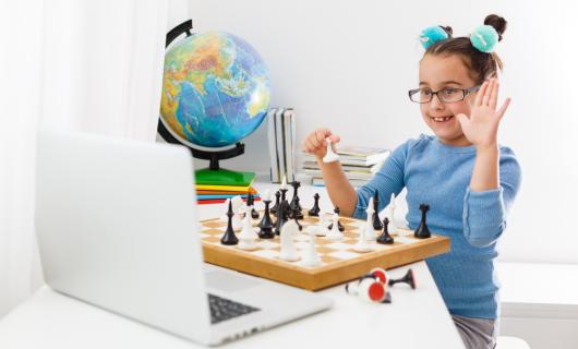 Fotografía de una niña jugando ajedrez frente a un computador, al fondo un mapamundi