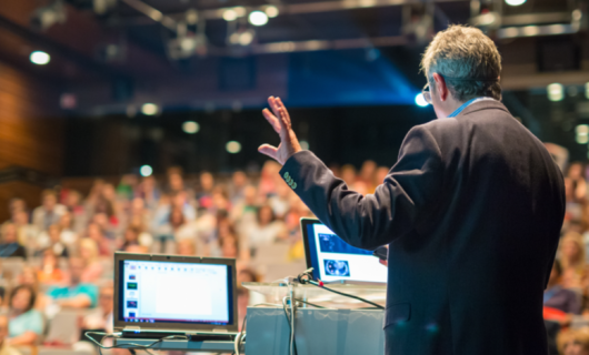 Conferencista de espaldas, hombre, en un auditorio lleno de personas, con su mano levantada, de pie y dos computadores de apoyo se dirige al público