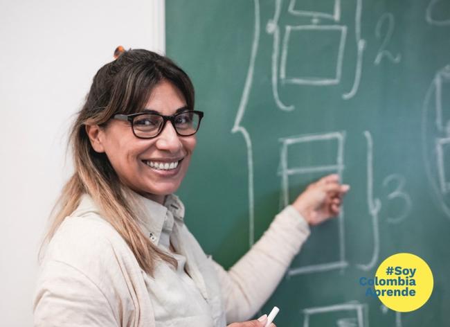 Foto de una profesora sonriendo y señalando un tablero