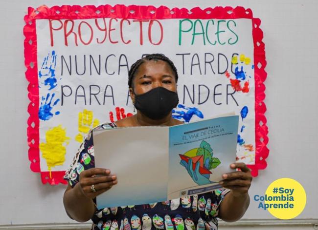 Foto de una mujer con tapabocas parada delante de una cartelera sosteniendo un cuadernillo de estudio